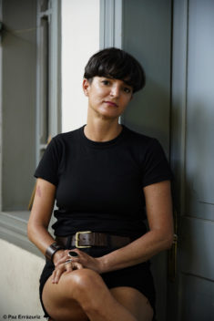 Ángeles, seated on a high stool in the center of the frame, is wearing a black shortsleeved shirt and dark jean shorts with a wide brown leather belt. She is wearing a leather bracelet and big rings, and her hands are clasped on her knee. Behind her is a white wall, a grey window frame, and a grey door.