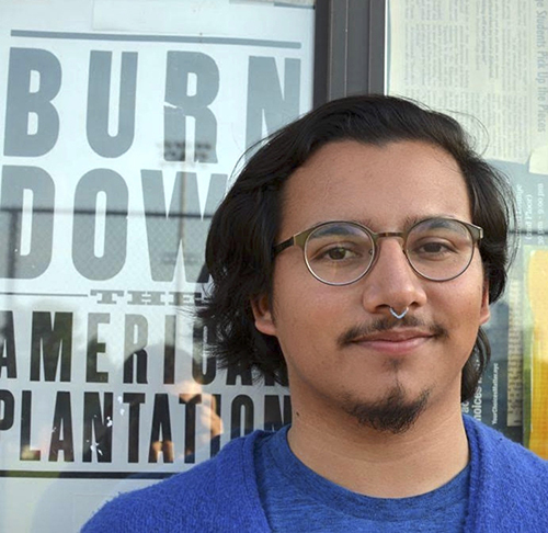 Photo Description: Light-skinned young man stares directly at the camera. His hair is short length, and he is wearing a cerulean blue shirt, topped with a cardigan of the same color. He wears glasses and had a septum piercing. He stands in front of a window that holds a sign that says “Burn Down the American Plantation.”