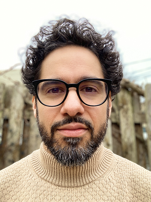 Photo Description Ricardo Gabriel is standing in front of a tall wooden fence and an open sky. He has light brown skin; wavy, medium-length black hair; and a short black beard with a touch of grey. He is wearing a light brown sweater and black-rimmed glasses.