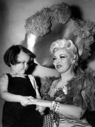 Mae West appears, with her platinum blonde hair and a large shiny hat, smiling at a young girl with a bob haircut and holding her hand.