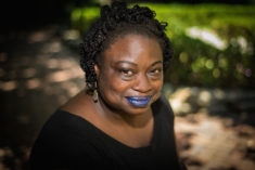 An image of a confident Black woman who gazes up toward the camera with a slight smile. Her blue lipstick stands out against her simple black attire. The background is out of focus but appears to be a pathway and a patch of grass shaded by trees.